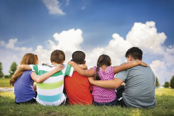group of children, arm in arm with one another. the children's backs are to the camera, and there's a blue sky with puffy clouds in the background.