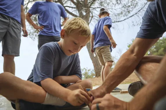Young child drawing on the ground with (out of shot) camp staff. He has a focused look on his face, not looking at the camera.