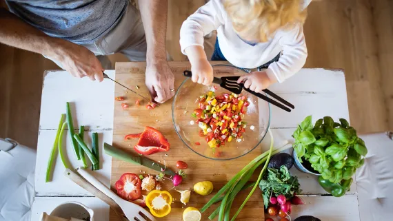 A cooking scene from above, you can see the hands of an adult and a child. The adult is chopping red bell peppers, the child is standing over the mixing bowl, assisting.